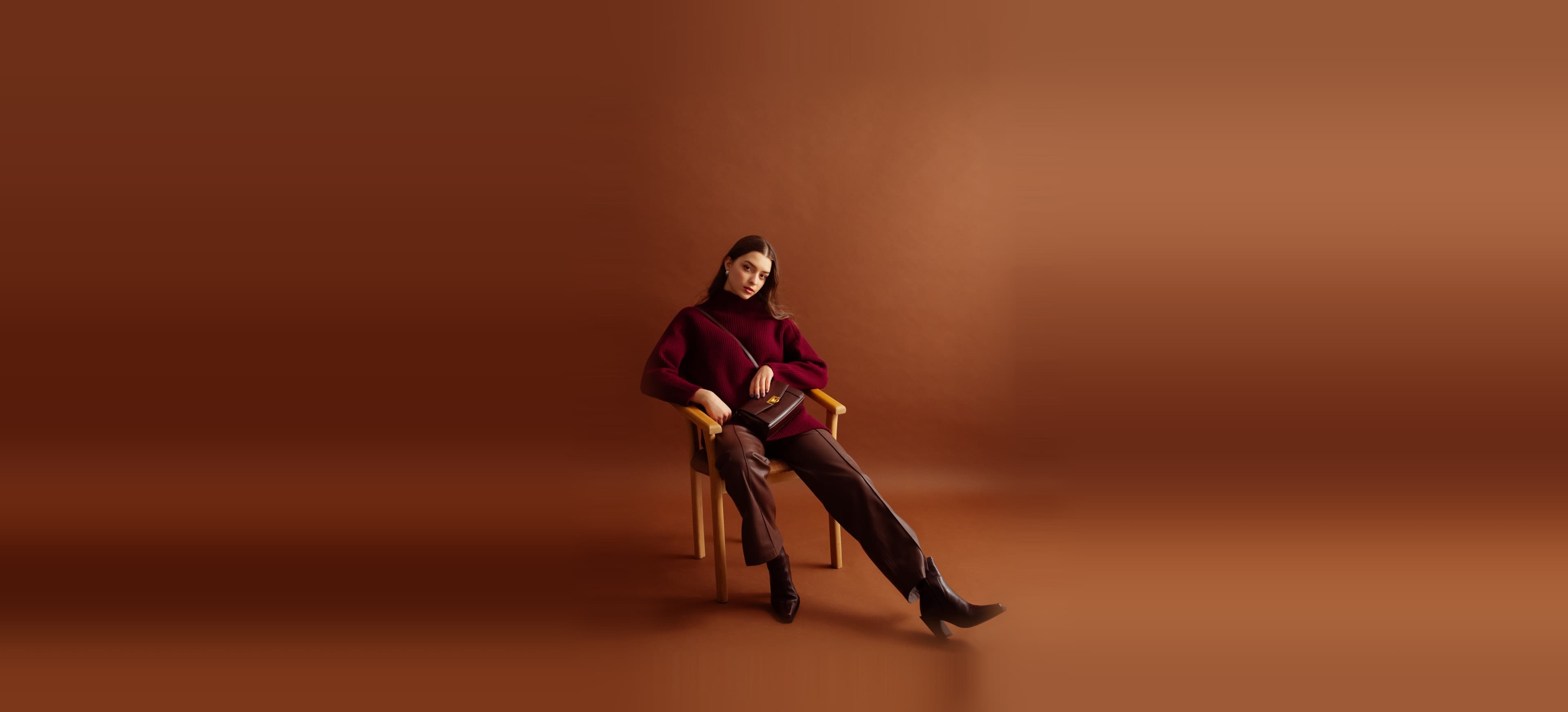 A woman in a burgundy turtleneck and brown leather trousers, seated in a wooden chair against a brown studio backdrop